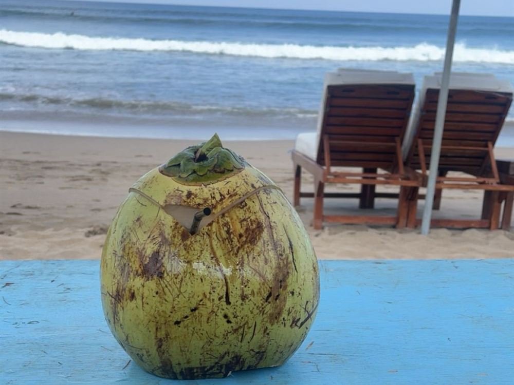 A fresh coconut on a blue table at the beach in Bali, with sun loungers and waves behind